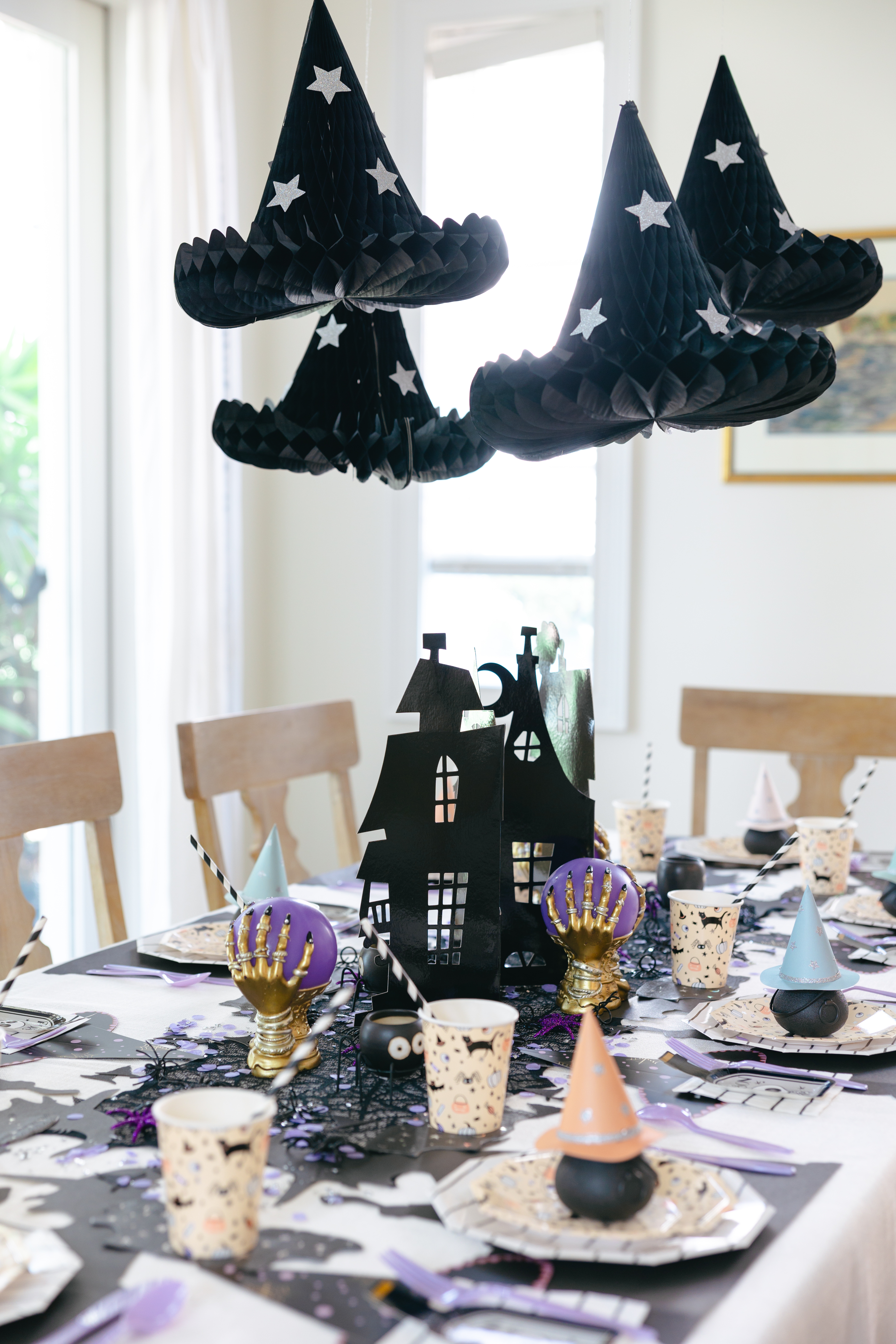 Table set for a party with pastel Halloween paper goods and honeycomb witch hats hanging overhead
